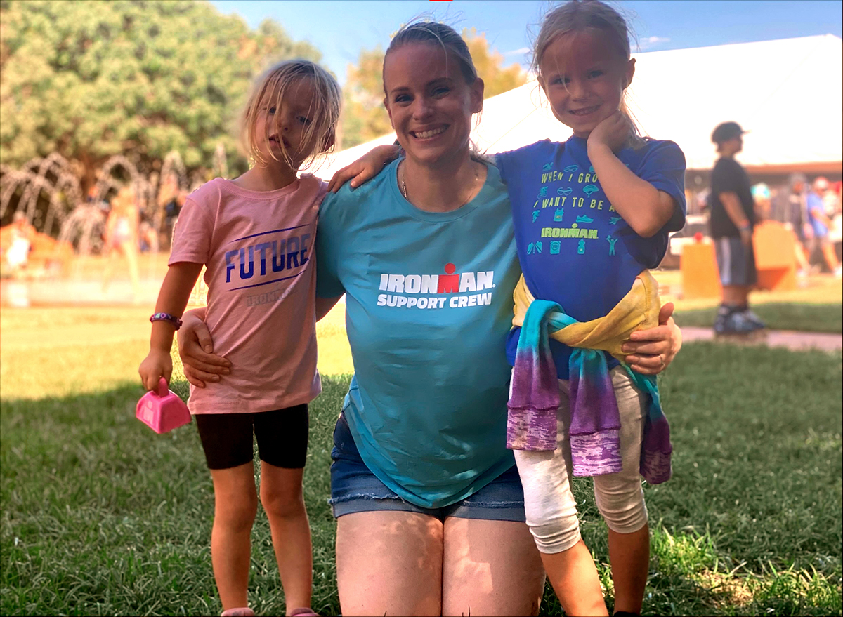 a smiling white woman with blond hair kneels on the grass at a triathlon with her arms around her two young daughters on either side. Her blue tshirt says "Ironman Support Crew." One daughter holds a pink bell in one hand and is wearing a pink tshirt that says "Future Ironman" and the other daughter has a multicolored tie-dyed sweatshirt tied around her waist and is wearing a blue tshirt that says "When i grow up i want to be an Ironman"