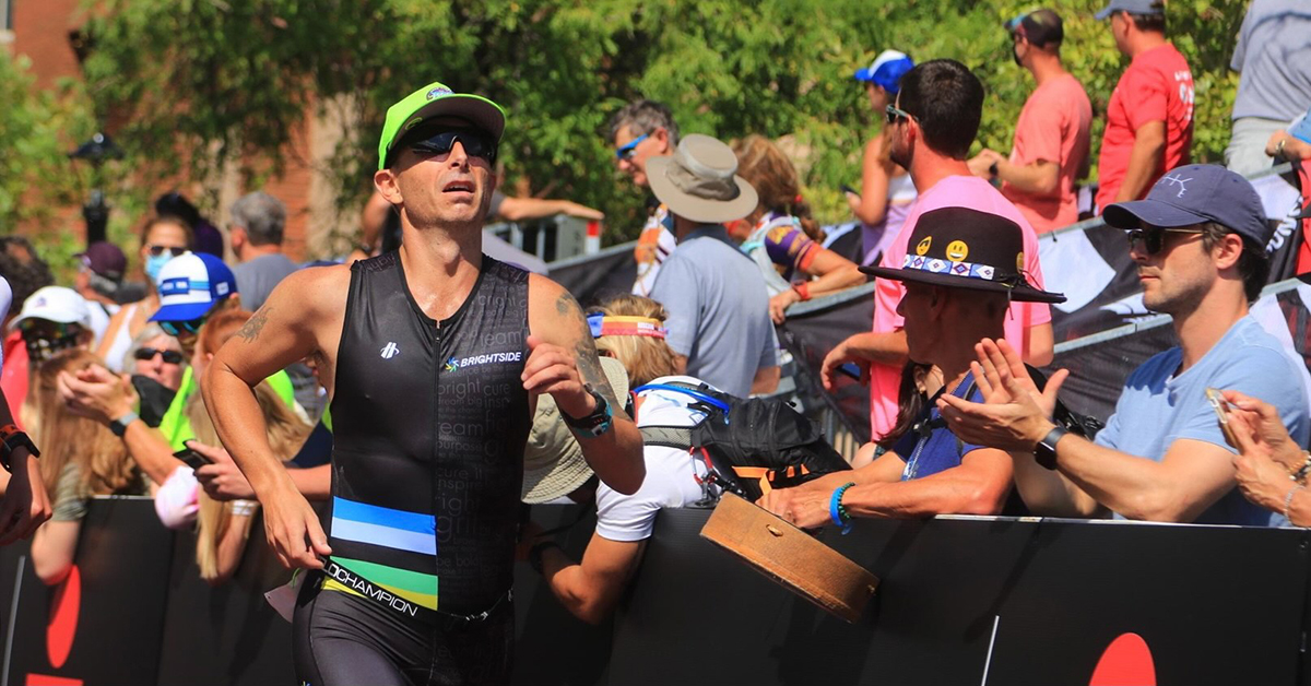 Brett Heyes, Credico's Director of National Accounts, competing in the 2021 Utah Ironman 70.3. He is seen running past an applauding crowd wearing a Team Bright Side tri kit, sunglasses, and a bright green cap.