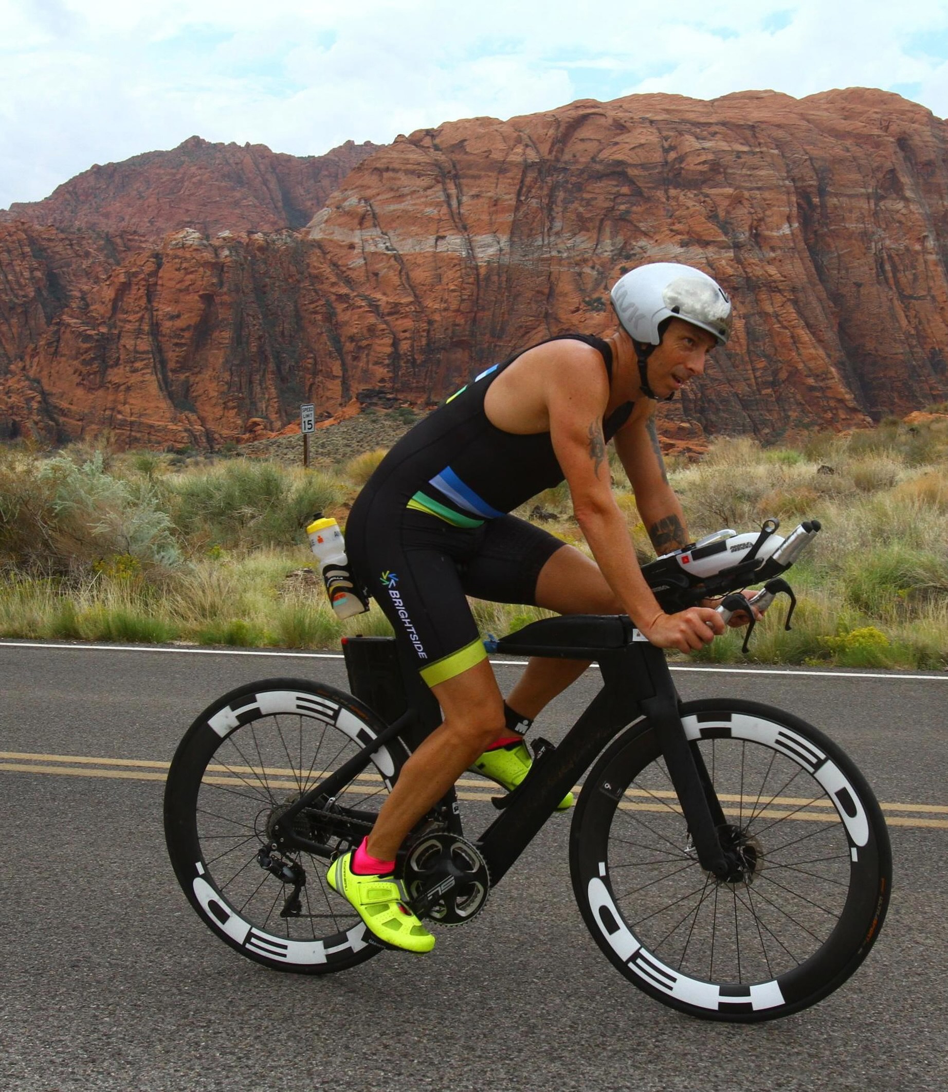 Brett Heyes, Credico's Director of National Accounts, rides past the red rocks of Utah. He is wearing a Team Bright Side tri kit, a silver helmet, and hi-viz yellow cycling shoes on a black bicycle with aero bars.