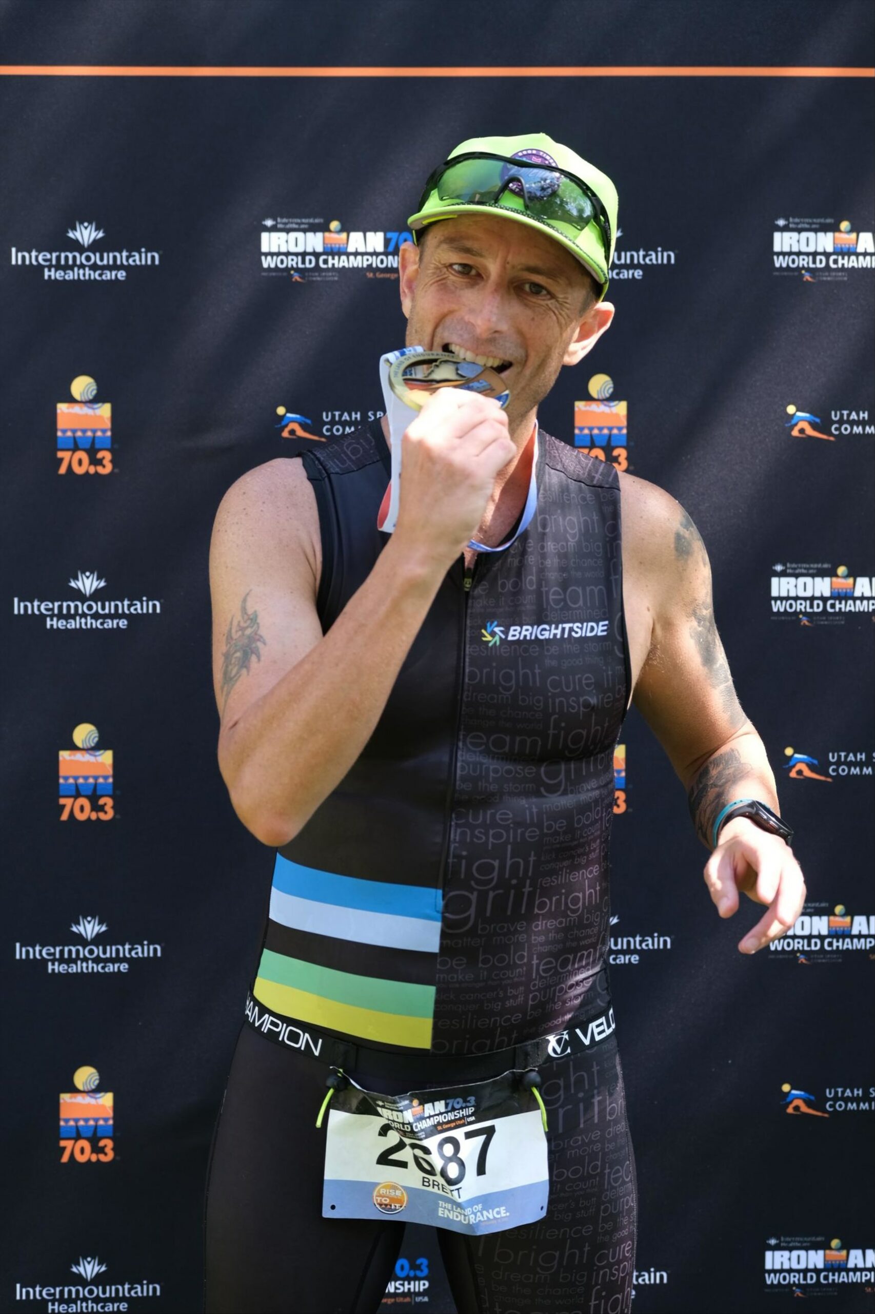 Brett Heyes, Credico's Director of National Accounts, bites his medal in front of an Ironman backdrop in his tri kit after completing a race.