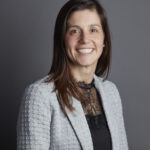 Josie Agro, a smiling white woman with long dark hair and a grey suit jacket over a black top