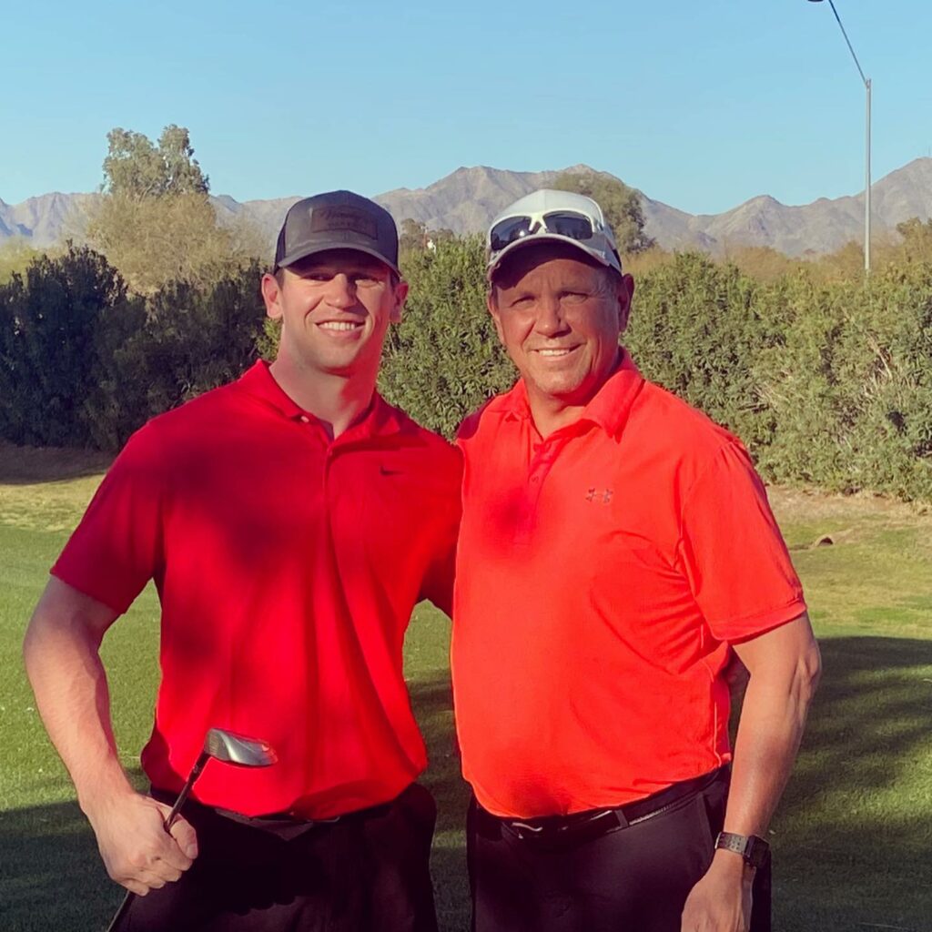 two men wearing hats and red polo shirts smile on the golf course holding clubs