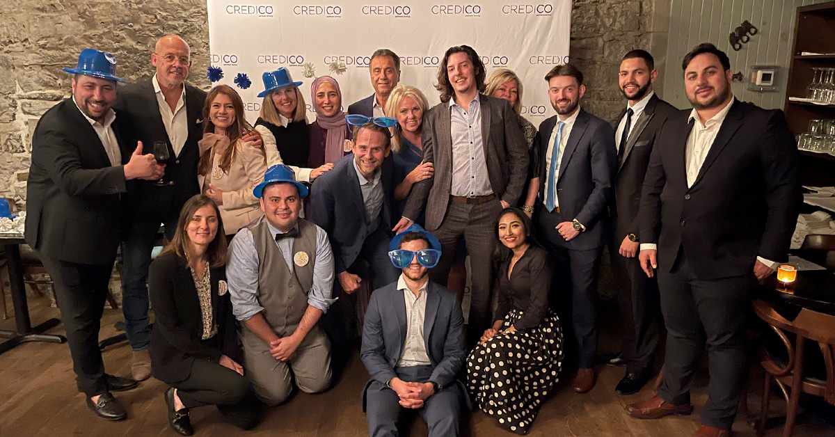 members of Credico's Canadian and US teams pose as a group in front of a Credico-branded backdrop wearing fun blue hats, glasses, pins, and other celebratory accessories