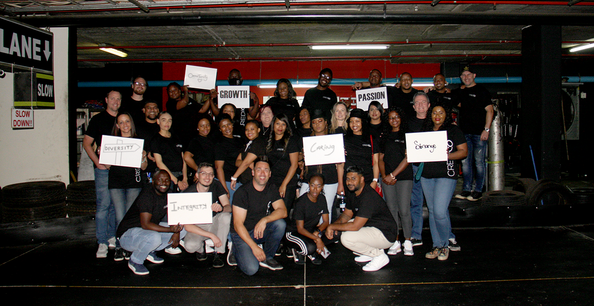 Credico South Africa poses for a group photo at their celebratory go-kart outing, holding signs displaying words that reflect some of our core values like integrity, diversity, opportunity, passion, growth, caring, and simunye, meaning "we are one"