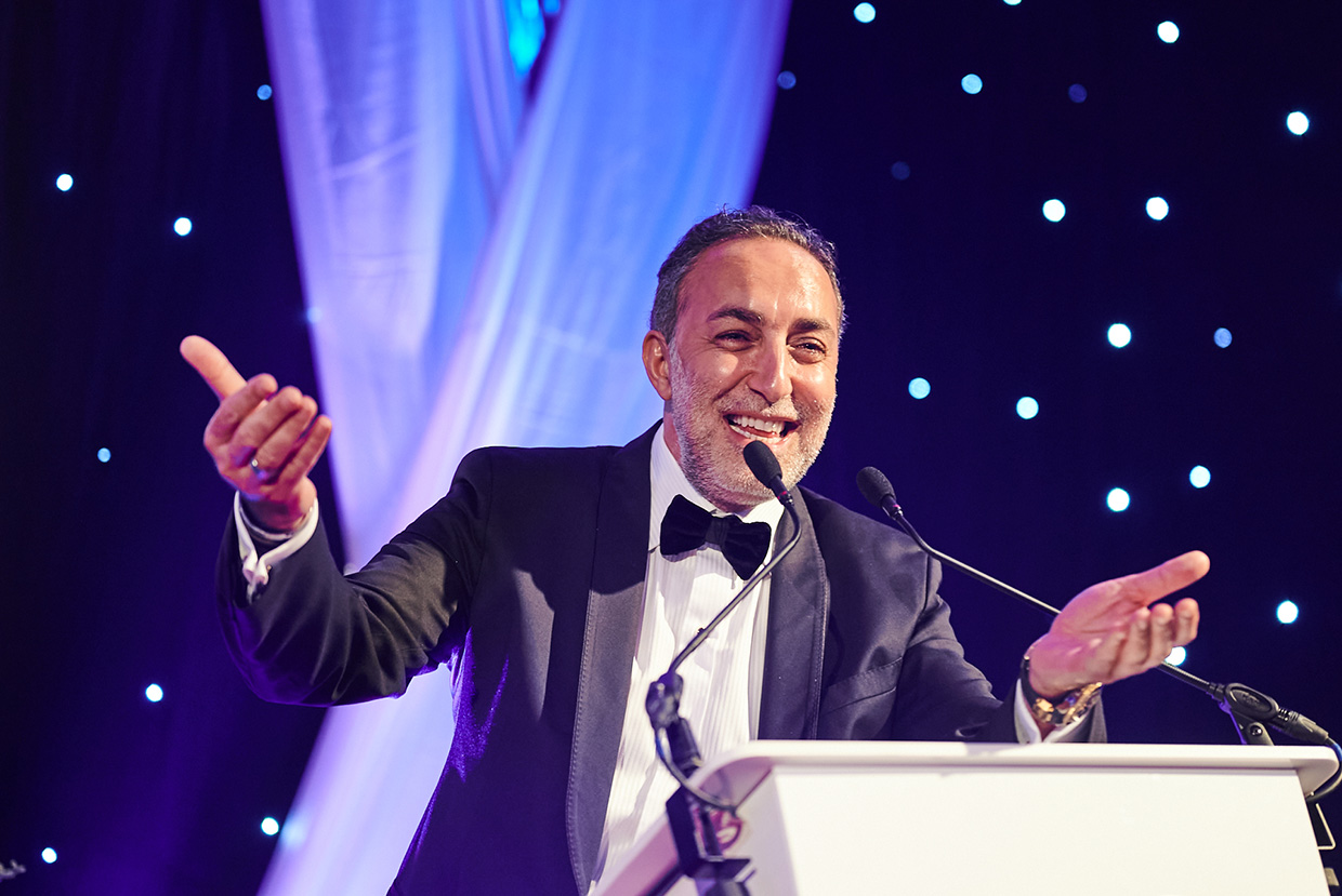 Credico's Chairman, Antoine Nohra, speaks from behind a podium on stage at the 2022 UK Business Awards in London. He is smiling and holding his hands out in a congratulatory gesture.