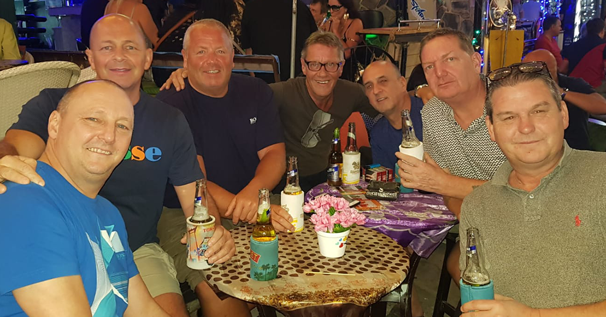 John Dougan with six friends, seated around a table with drinks at a tropical-themed venue