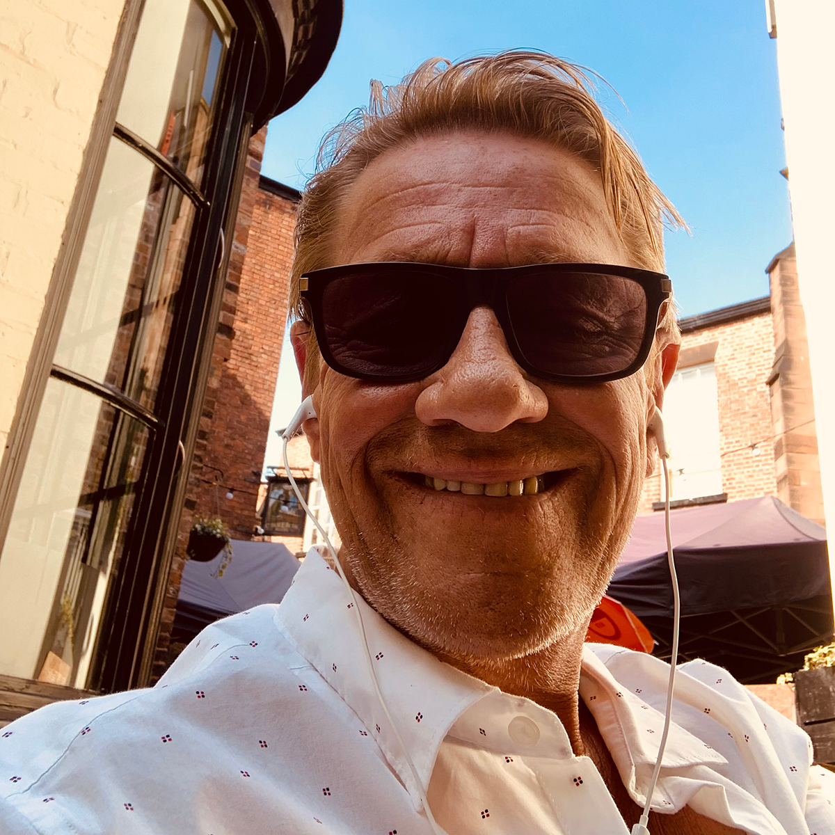 John Dougan smiles in a selfie taken outdoors. He is wearing sunglasses and has white corded earbuds in his ears.