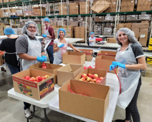 Credico team members wearing aprons, gloves, and hairnets pause to smile for a photo while packing apples at a volunteer event at the Greater Chicago Food Depository. From left to right: Devin Gomez, Onboarding Specialist; Michael Fergus, Assistant Commissions Manager; Lea Marrs, Assistant Corporate Counsel, Brennan Delaney, Commissions Analyst (seated in the background); Jen Goldfarb, Director of Human Resources.
