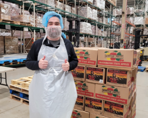 Conor Brennan, Senior Accountant at Credico USA, smiles from behind a beard net with a thumbs-up gesture in front of a pallet of cases of apples during a volunteer day at the Greater Chicago Food Depository