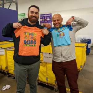 Two Credico volunteers hold up children's Crayola shirts that say "Super Cool Dude" and "Creative Dude" while sorting clothing at Cradles to Crayons