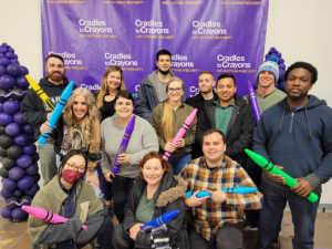 Part of the Credico team poses as a group in front of a Cradles to Crayons backdrop while smiling and holding large plastic crayons.