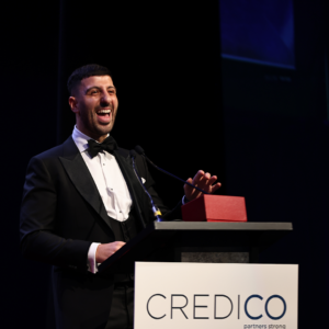 Chris Mikkides, wearing a tuxedo, gives a speech on stage behind a podium following his recognition as a Regional Consultant at the January 2023 North American Awards Gala in Miami, Florida