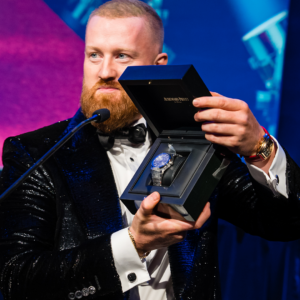 Gavin Walsh, wearing a sparkling black tuxedo, holds up an Audemars Piguet watch in a case while on stage behind a podium following his recognition as a National Consultant at the January 2023 North American Awards Gala in Miami, Florida