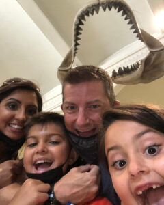 Jeremy Klarner and his family smile with wide open mouths in a selfie taken at a museum under a large set of shark jaws