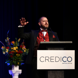 James Flynn, wearing a tuxedo with a red vest, gives a speech on stage behind a podium following his recognition as a Regional Consultant at the January 2023 North American Awards Gala in Miami, Florida