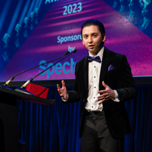 Michael Wang, wearing a tuxedo, gestures while giving a speech on stage behind a podium following his recognition as a Regional Consultant at the January 2023 North American Awards Gala in Miami, Florida