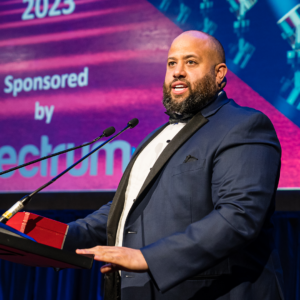 Obiora Shorinwa, wearing a navy tuxedo with black lapels, gives a speech on stage behind a podium following his recognition as a Regional Consultant at the January 2023 North American Awards Gala in Miami, Florida