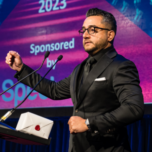 Pani Verma, wearing a black suit, shirt, and tie, gestures while giving a speech on stage behind a podium following his recognition as a Regional Consultant at the January 2023 North American Awards Gala in Miami, Florida