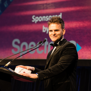 Ronan O'Connor, wearing a tuxedo, gives a speech on stage behind a podium following his recognition as a Regional Consultant at the January 2023 North American Awards Gala in Miami, Florida