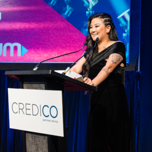 Simone Suetsugu, wearing a floor-length black velvet dress, gives a speech on stage behind a podium following her recognition as a Regional Consultant at the January 2023 North American Awards Gala in Miami, Florida