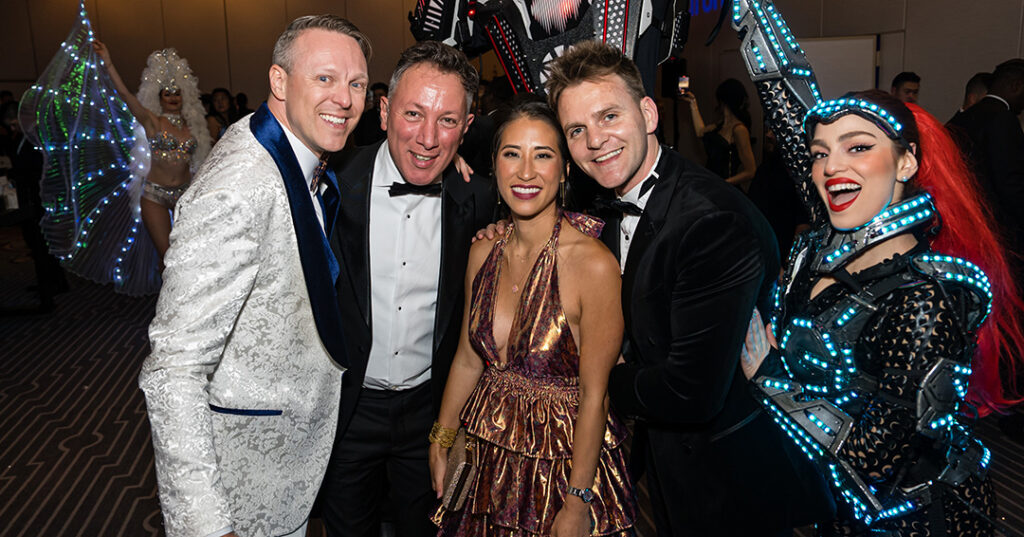 A group of attendees dressed in glittering formalwear pose for a photo with costumed entertainers during the January 2023 North American Awards Gala in Miami, Florida