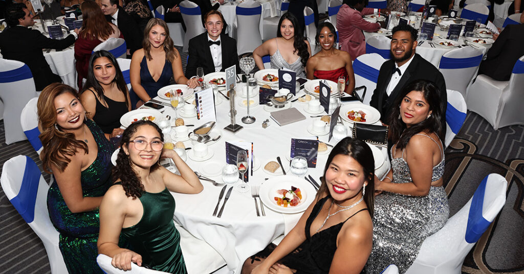 A group of attendees dressed in glittering formalwear smile from their seats around a table at the January 2023 North American Awards Gala in Miami, Florida