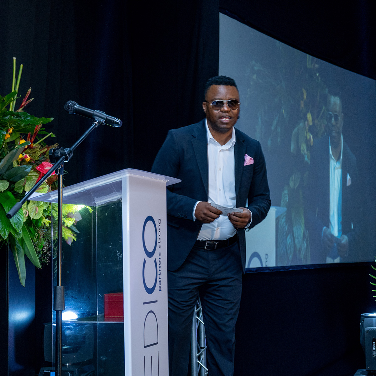 Kevin de Souza, wearing sunglasses on stage, addresses the audience from next to a podium with his image projected on-screen next to him at Credico South Africa's 2023 Awards Gala at Sun City Resort